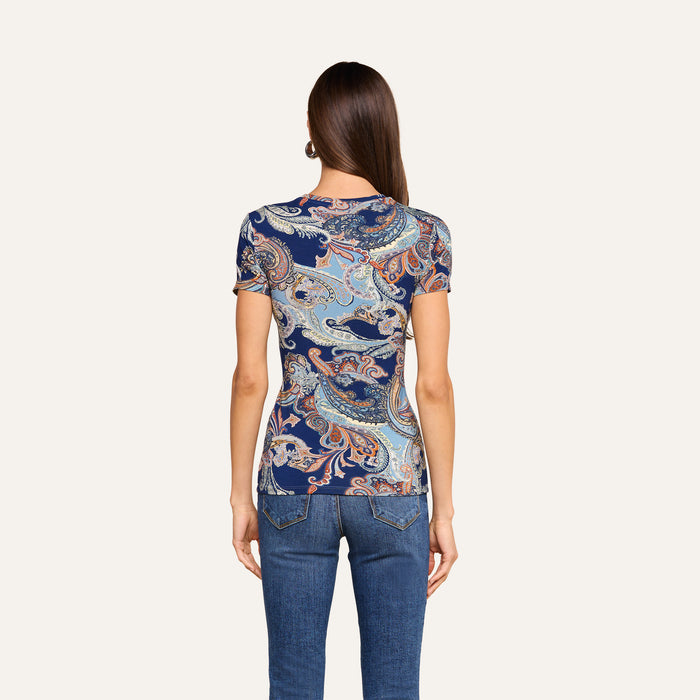 A woman with long brown hair, wearing the Ressi Fitted Tee in midnight blue paisley and blue jeans, is shown from the back against a plain light background.