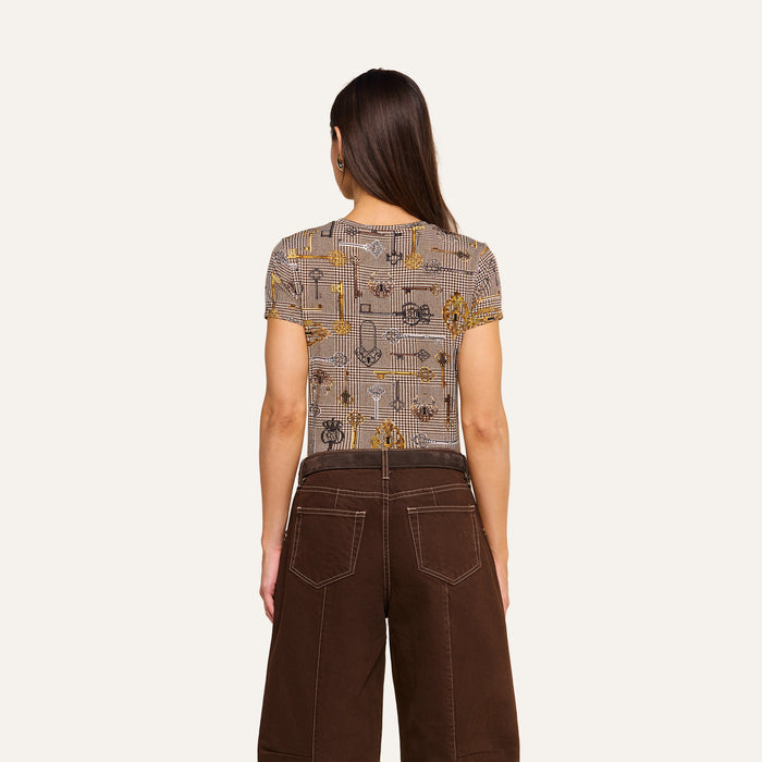 A woman with long brown hair stands facing away, wearing the bestselling Ressi Fitted Tee and high-waisted brown pants with white stitching, against a plain light background.