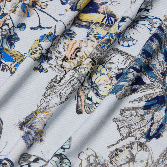 Close-up of the Ressi Fitted Tee’s stretch jersey knit fabric, showcasing a blue butterfly print complemented by floral designs in shades of yellow and beige. The material drapes in soft folds, casting gentle shadows and highlights across the surface.