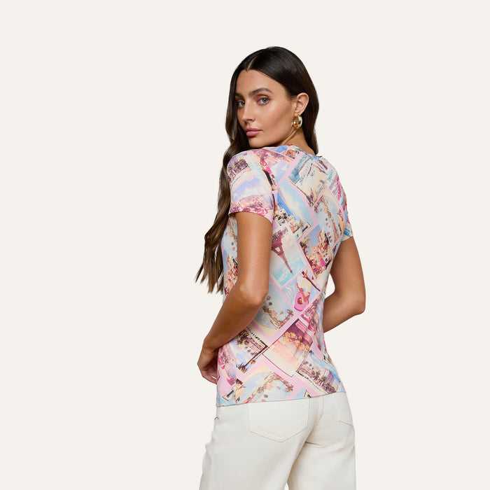 A woman with long brown hair poses with her back turned, looking over her shoulder against a plain, light background. She is wearing the bestselling Ressi Fitted Tee—a colorful short-sleeve top in stretch jersey knit—paired with white pants.