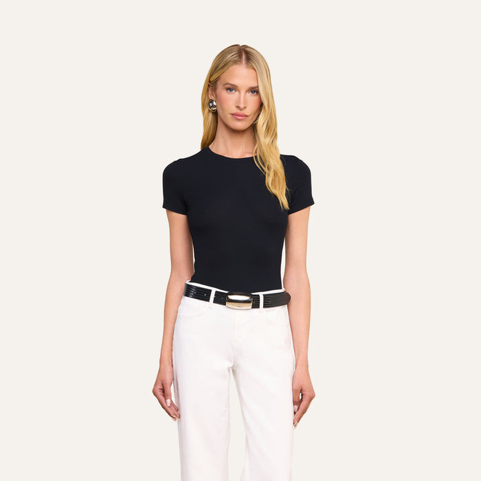 A woman with long blonde hair wears the black Ressi Micro-Rib Tee, paired with white pants, a black belt featuring a large gold buckle, and earrings, standing against a plain light background.