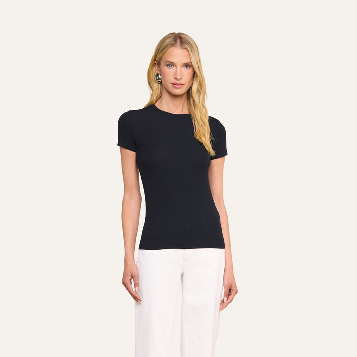 A person with long blonde hair wears the slim-fit, short-sleeve black Ressi Micro-Rib Tee and white pants, standing against a plain light background.