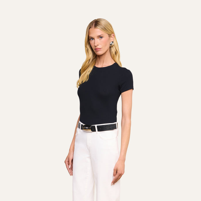 A woman stands against a plain background wearing the black Ressi Micro-Rib Tee styled with white pants, a black belt, and large earrings. She has long blonde hair and a neutral expression.