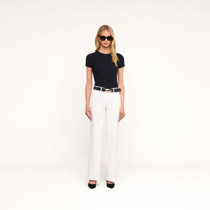 A woman with long blonde hair wears sunglasses and a black Ressi Micro-Rib Tee, paired with white high-waisted pants, a black belt, and black heels, standing against a plain white background.