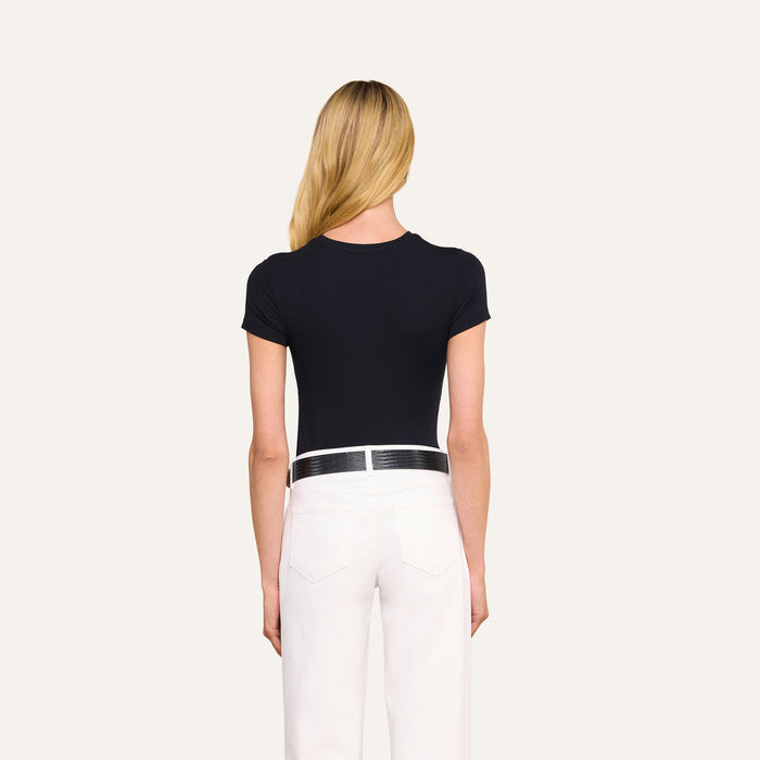 A person with long blonde hair stands facing away, wearing the black Ressi Micro-Rib Tee, white pants, and a black belt against a plain light background.