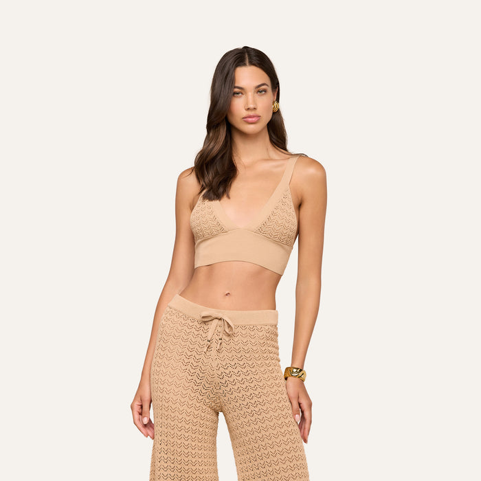 A woman with wavy brown hair wears the Reverie Pointelle Knit Bralette in beige, featuring a deep V-neckline, paired with matching high-waisted drawstring pants, standing against a plain cream background.