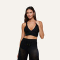 A woman wearing the Reverie Pointelle Knit Bralette, a black top with a deep V-neckline, paired with matching high-waisted drawstring pants featuring a lacy pointelle pattern, stands against an off-white background and poses with one hand on her strap.