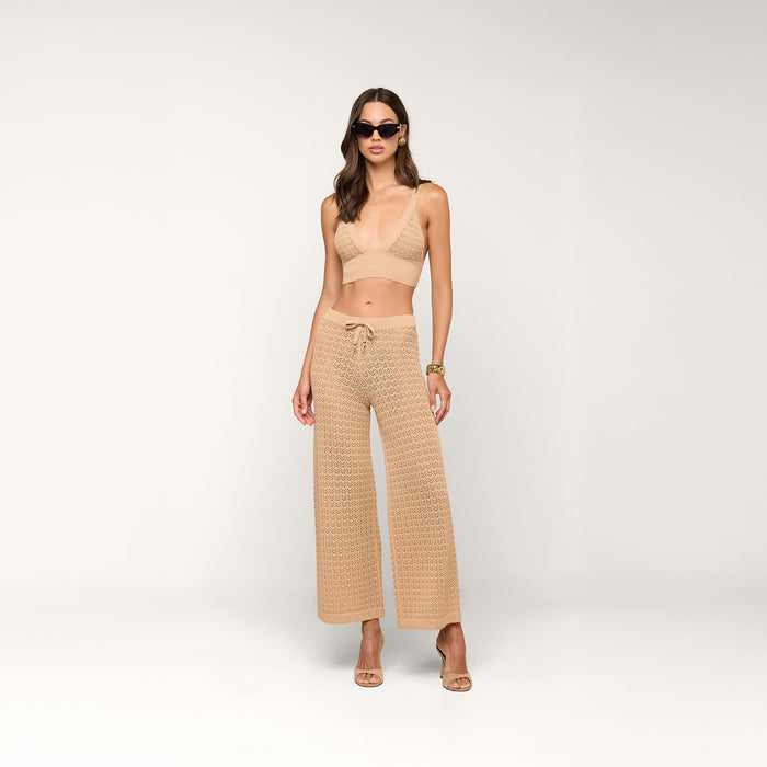 A woman wearing sunglasses, the Reverie Pointelle Knit Bralette in beige with a deep V-neckline, along with matching wide-leg pointelle knit pants featuring a tie at the waist, and high-heeled sandals, standing against a plain white background.