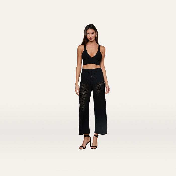 A woman stands against a plain background wearing the Reverie Pointelle Knit Bralette, which features a black hue and deep V-neckline. She pairs it with black crochet pants and black heeled sandals. Her long hair is down, and she poses in a relaxed stance with her arms at her sides.