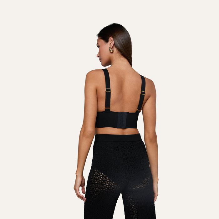 A woman stands facing away, wearing the Reverie Pointelle Knit Bralette with thick straps and high-waisted black lace pants. Her hair is down, and she wears hoop earrings, set against a plain, light background.