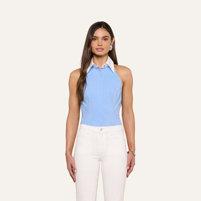 A woman stands against a plain light background, wearing the Rina Poplin Halter Top—a sleeveless, collared light blue blouse made from organic cotton—paired with high-waisted white pants. Her long brown hair is styled straight.