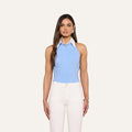 A woman stands against a plain light background, wearing the Rina Poplin Halter Top—a sleeveless, collared light blue blouse made from organic cotton—paired with high-waisted white pants. Her long brown hair is styled straight.
