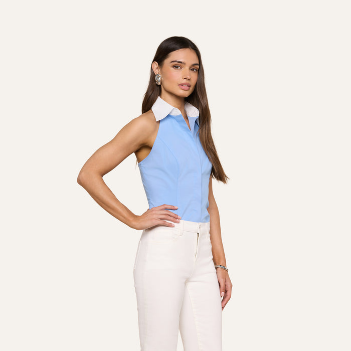 A woman stands against a plain background, wearing the Rina Poplin Halter Top in light blue organic cotton paired with white pants. She has long straight hair and accessorizes with earrings and a bracelet.