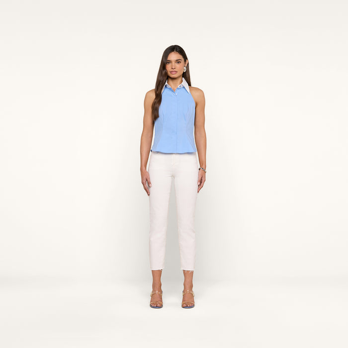 Woman with long brown hair stands facing forward, wearing the Rina Poplin Halter Top—a sleeveless light blue summer blouse—paired with white pants and beige heeled sandals, against a plain white background.