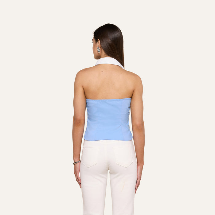 A woman stands with her back to the camera, wearing the light blue Rina Poplin Halter Top and white pants against an off-white background.
