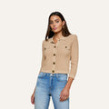 A woman with long brown hair wears the Risa Collared Cardigan—a beige, button-up, pointelle knit sweater with two chest pockets—paired with blue jeans, standing against a plain off-white background.