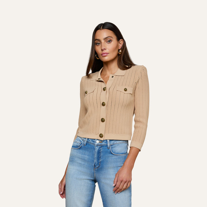 A woman with long brown hair wears the Risa Collared Cardigan—a beige, button-up, pointelle knit sweater with two chest pockets—paired with blue jeans, standing against a plain off-white background.