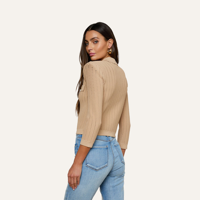 A woman stands sideways and looks over her shoulder, wearing the Risa Collared Cardigan—a beige pointelle knit sweater with three-quarter sleeves—paired with light blue jeans, against a plain light background.