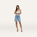A woman stands with arms crossed, wearing a white tank top, beige heels, and the River Jean Bermuda Short—an all-cotton, mid-rise denim Bermuda short—against a plain light background.