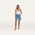 A woman stands against a plain white background wearing a white tank top, nude heels, and the River Jean Bermuda Short— a mid-rise denim Bermuda short made from stretch denim.