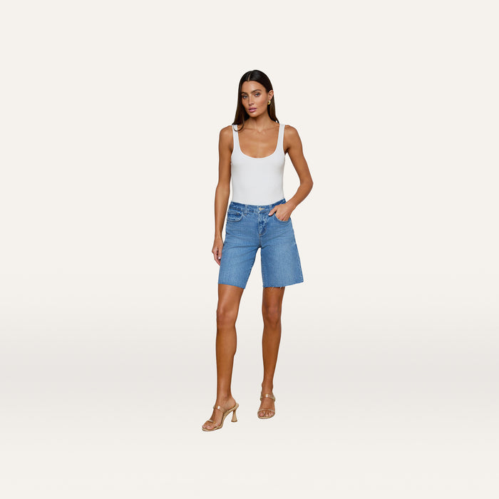 A woman stands against a plain white background wearing a white tank top, nude heels, and the River Jean Bermuda Short— a mid-rise denim Bermuda short made from stretch denim.