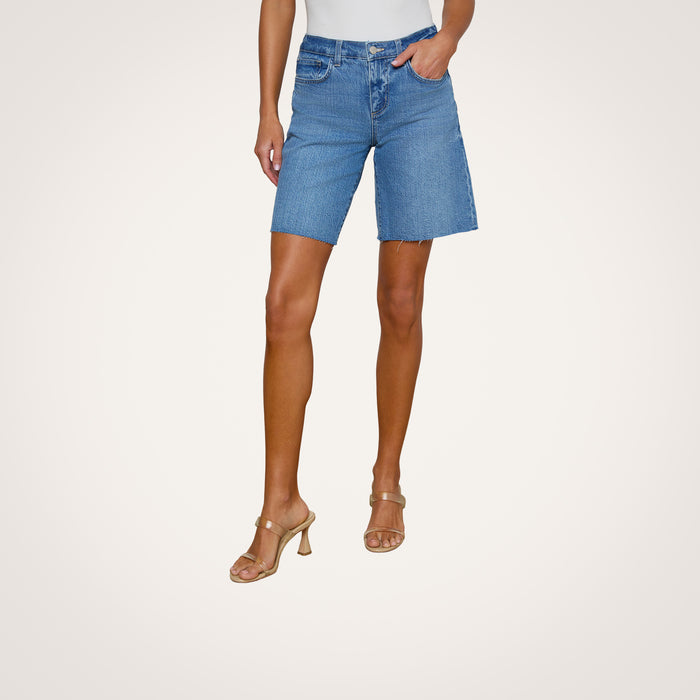 A person wearing a white top, the River Jean Bermuda Short in mid rise blue stretch denim, and beige heeled sandals poses with one hand in a pocket against a plain light background.
