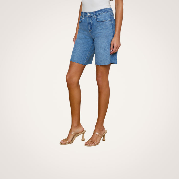 A woman wearing the River Jean Bermuda Short in blue stretch denim, paired with a white top and beige heeled sandals, stands against a plain, light background with her arms relaxed by her sides.