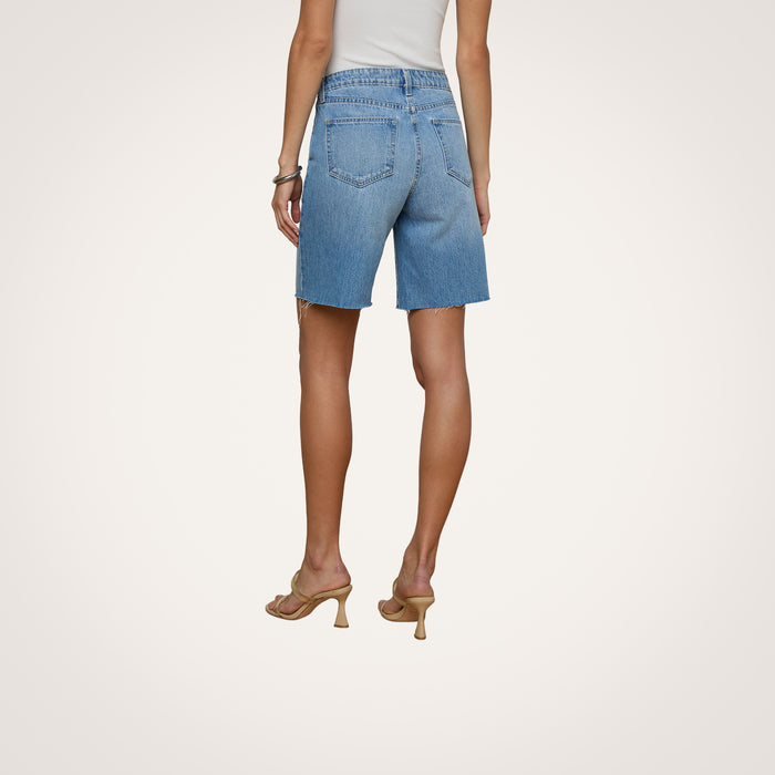 Wearing the River Jean Bermuda Short, a person in a white sleeveless top and beige heeled sandals stands with their back to the camera against a plain light background.