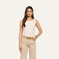 A woman with long dark hair is wearing the Riza Boatneck Bodysuit—a sleeveless, vintage-inspired white one-piece—paired with beige pants, a wide gold belt, and large gold bracelets and earrings, standing against a plain off-white background.