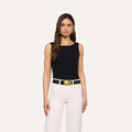 Woman with long brown hair wearing the sleeveless L’AGENCE Riza Boatneck Bodysuit, paired with white pants and a white belt featuring a large gold buckle, standing against a plain light background.