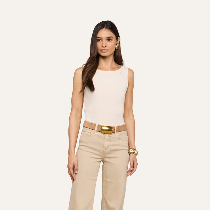 A woman with long brown hair wears a sleeveless beige top, beige pants, a gold belt, gold earrings, and a gold bracelet—her outfit complemented by the vintage-inspired Riza Boatneck Bodysuit in white—as she stands against a plain light background.