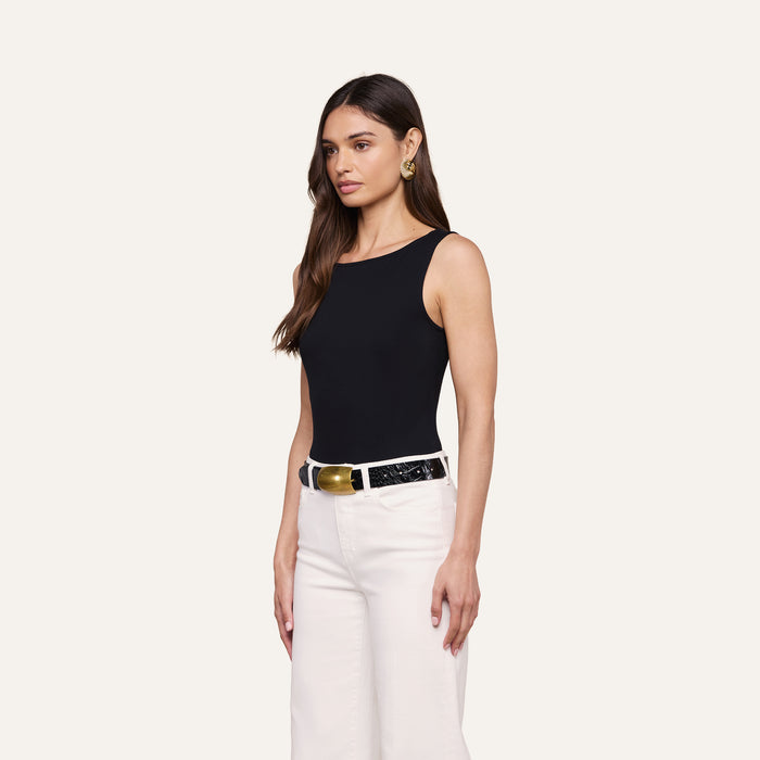 A woman stands against a plain background wearing the Riza Boatneck Bodysuit in black, paired with white pants, a wide black belt, and gold earrings. She has long brown hair and a neutral expression, looking slightly to the side.