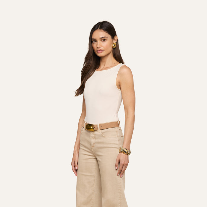 Woman with long brown hair wearing the Riza Boatneck Bodysuit in beige, styled with a light brown belt and beige wide-leg pants, accessorized with large gold earrings and bracelets, standing against a plain background.