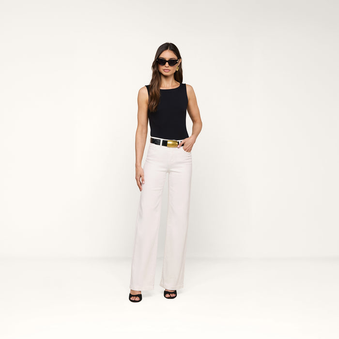 Wearing sunglasses and the Riza Boatneck Bodysuit in black, paired with white wide-leg pants, a black belt featuring a gold buckle, and black heels, a woman stands against a plain white background.