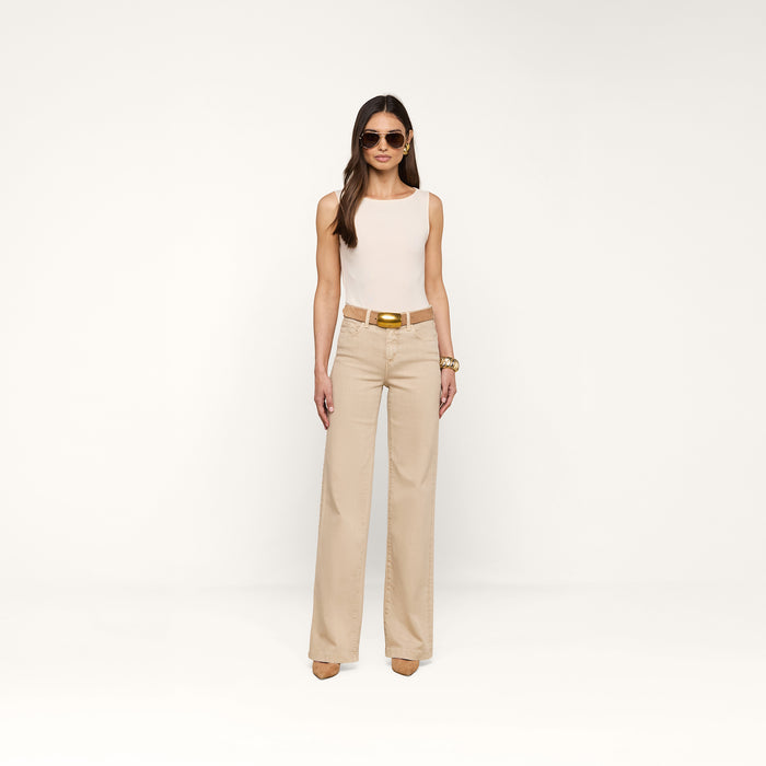 A woman wearing sunglasses, the Riza Boatneck Bodysuit in vintage white with a sleeveless design, beige wide-leg pants, a brown belt, and heels stands facing forward against a plain white background.