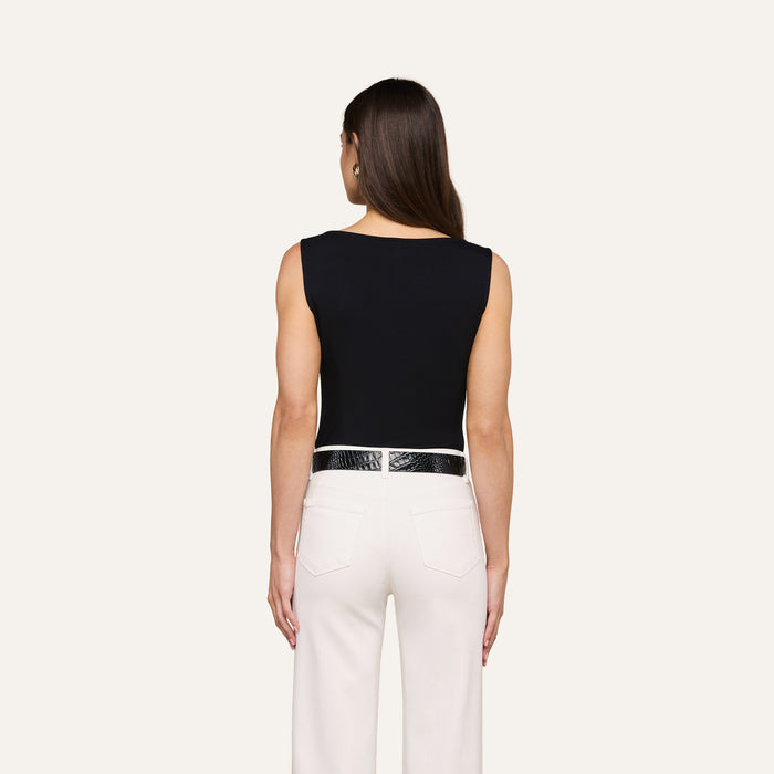 A woman stands facing away, wearing a sleeveless black Riza Boatneck Bodysuit, white pants, and a black belt. Her brown hair is straight and loose, and her arms are at her sides against a plain light background.