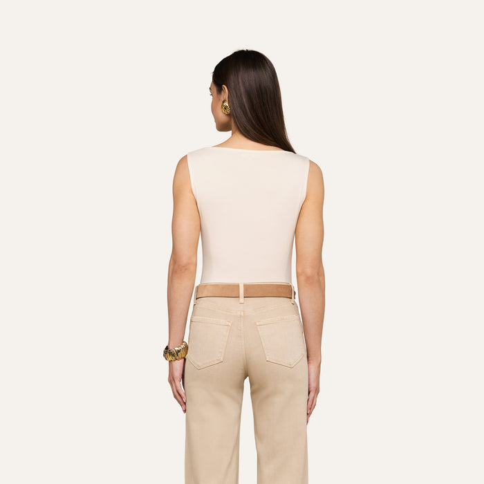 A woman stands with her back to the camera, wearing the sleeveless vintage white Riza Boatneck Bodysuit paired with beige jeans and a brown belt, accessorized with gold jewelry, against a plain light background.