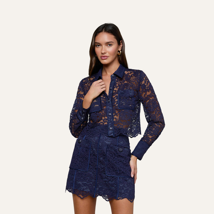 A woman wearing the Roelle Lace Blouse—a sheer navy lace top with scalloped edges—paired with a matching skirt featuring front pockets, stands against a plain light background.
