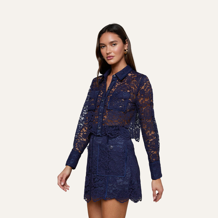 A woman wears the Roelle Lace Blouse in sheer navy with long sleeves, styled with a coordinating lace mini skirt. The ensemble is detailed with scalloped edges and patch pockets, set against a plain off-white background.
