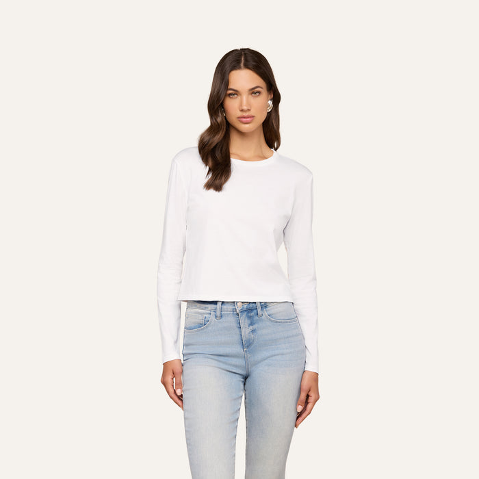 A woman with long brown hair wears the Rojene Cotton Tee, a relaxed fit plain white long sleeve shirt, paired with light blue jeans and standing against a plain light background.