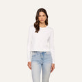 A woman with long brown hair wears the Rojene Cotton Tee, a relaxed fit plain white long sleeve shirt, paired with light blue jeans and standing against a plain light background.