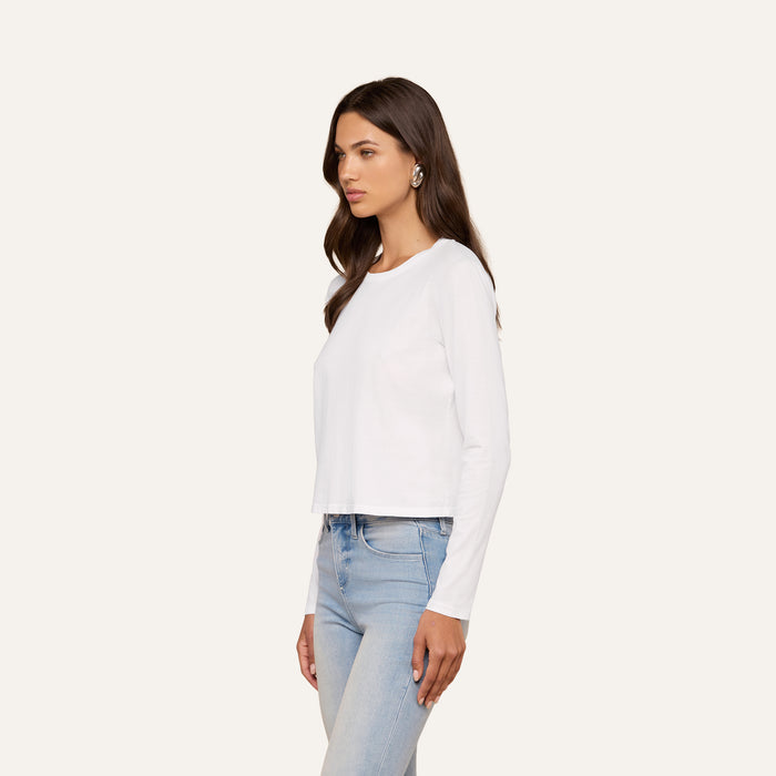 A woman with long brown hair wearing a relaxed fit, white Rojene Cotton Tee and light blue jeans stands facing sideways against a plain, light background.