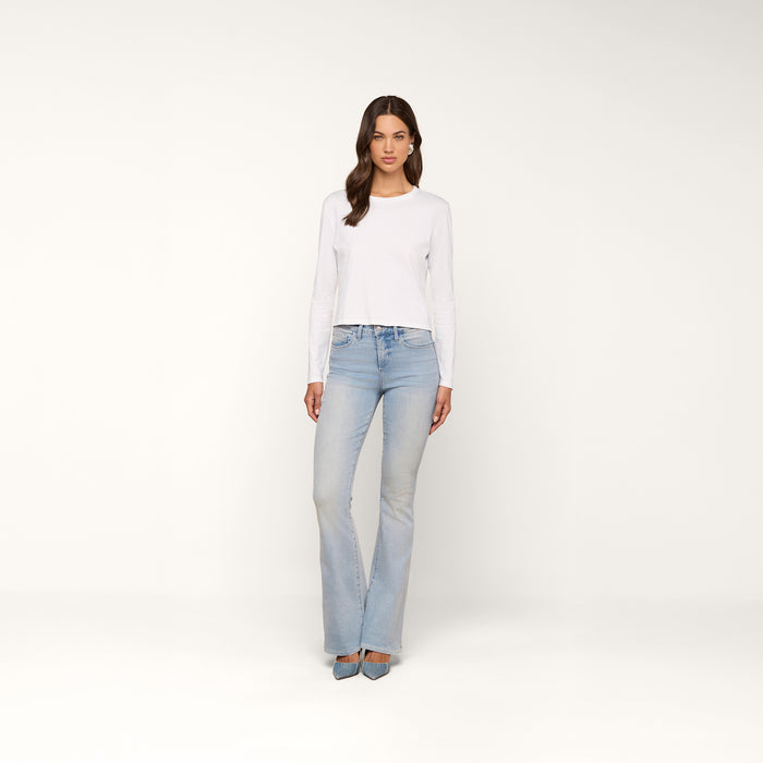 A woman with wavy brown hair and a neutral expression stands against a plain background, wearing the relaxed fit Rojene Cotton Tee paired with light blue flared jeans and pointed-toe shoes.