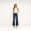 A woman stands against a white background wearing a sleeveless white halter top, the Rosalyn Wide-Leg Jean in stretch denim, a tan belt, and tan heels. She has long brown hair and one hand in her pocket.