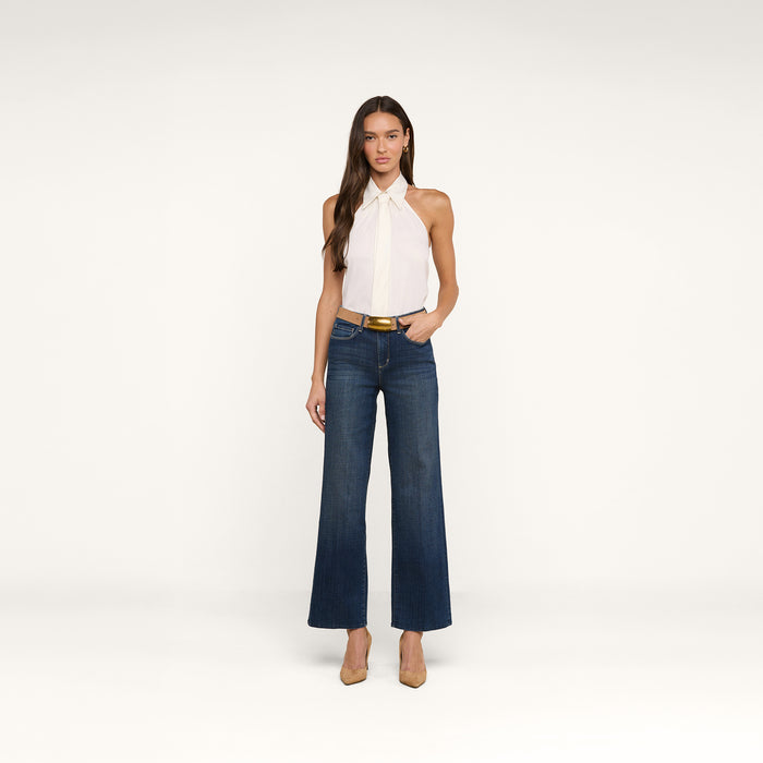 A woman stands against a white background wearing a sleeveless white halter top, the Rosalyn Wide-Leg Jean in stretch denim, a tan belt, and tan heels. She has long brown hair and one hand in her pocket.