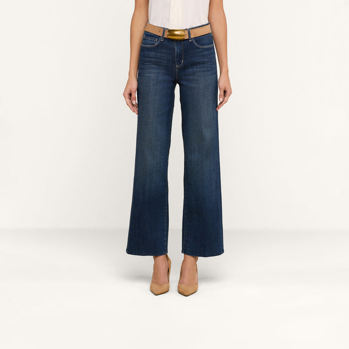 A person wearing a white top, the Rosalyn Wide-Leg Jean in high-rise stretch blue denim, a brown belt, and beige pointed-toe heels stands against a plain white background.