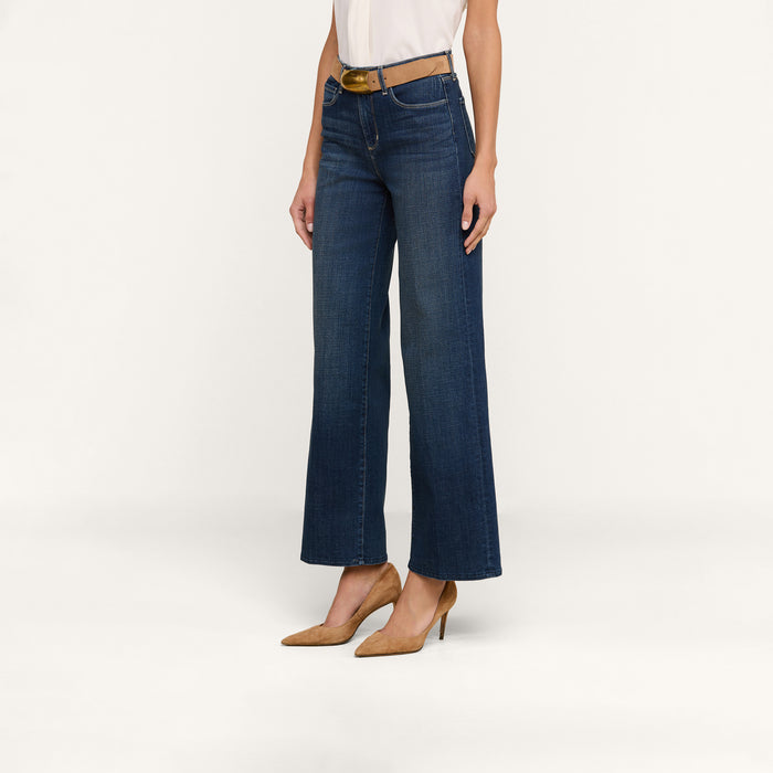 A person wearing the Rosalyn Wide-Leg Jean in blue stretch denim, paired with a white sleeveless top and tan high heels, stands against a plain white background.