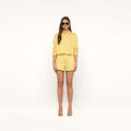 A woman wearing sunglasses and the Cosette Shirt, a yellow and white vertical striped tailored shirt with sleeves rolled up, paired with matching shorts and nude high-heeled sandals, stands against a plain white background.