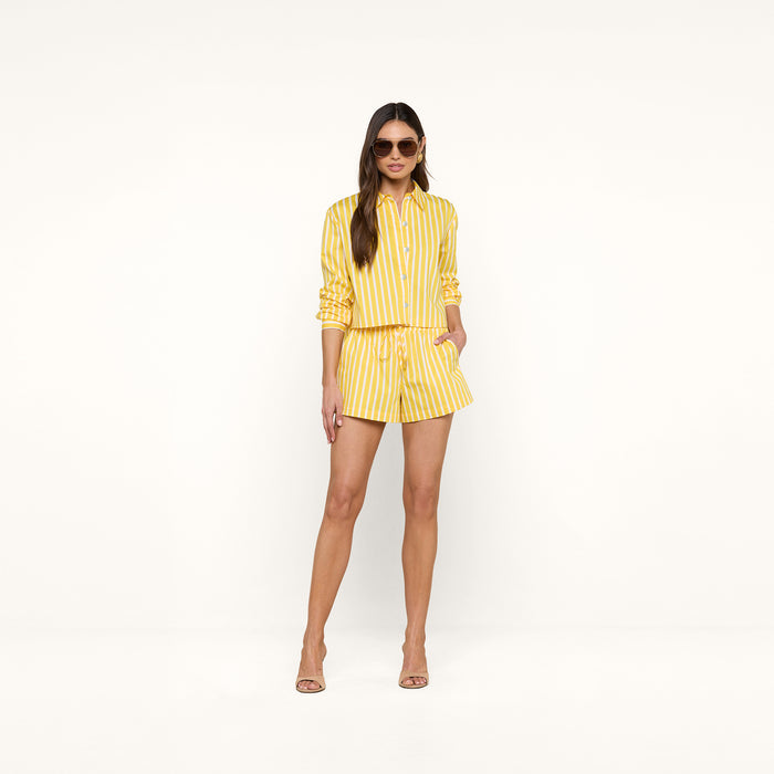 A woman wearing sunglasses and the Cosette Shirt—an organic cotton, yellow-and-white striped, long-sleeve top—paired with matching shorts stands against a plain white background with her hands in her pockets.