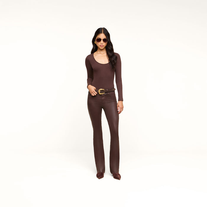 A woman wearing sunglasses, a long-sleeve brown top, and pointed shoes stands against a plain white background, looking at the camera with her hand on her belt. She completes the look with the Ruth Coated Straight-Leg Jean, adding a modern edge to her ensemble.